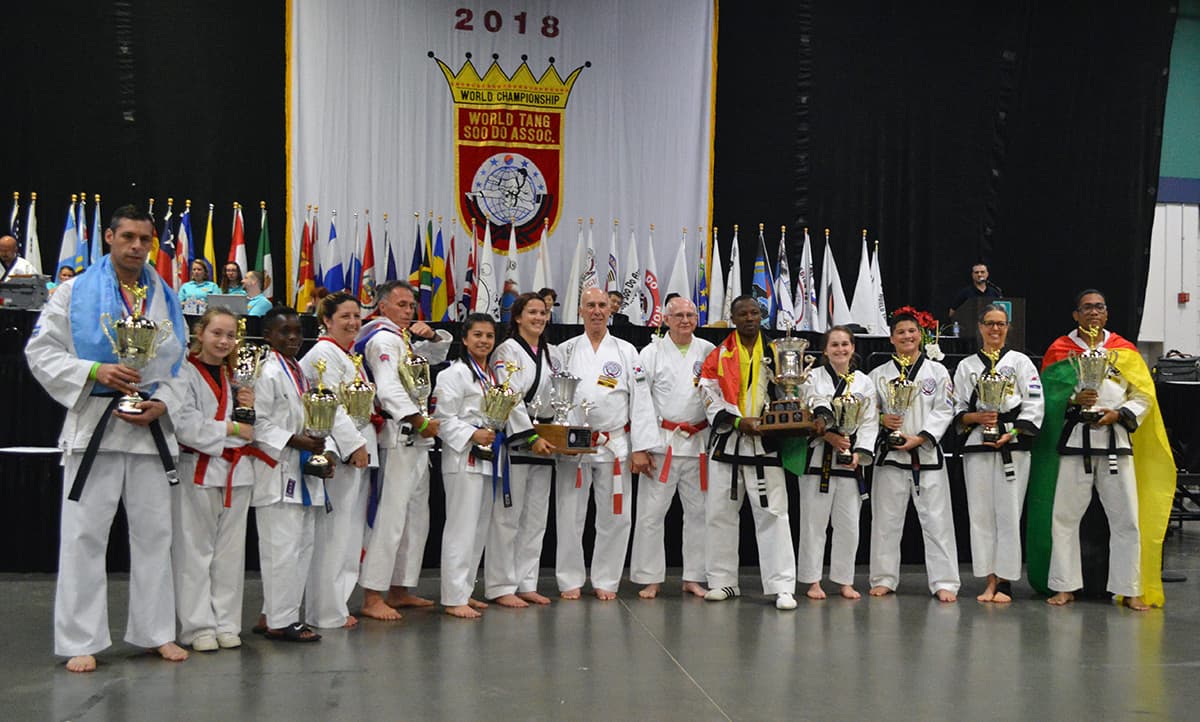 A lineup of Division Cup winners from many countries holding their trophies on stage in front of the World Championship flag backdrop.