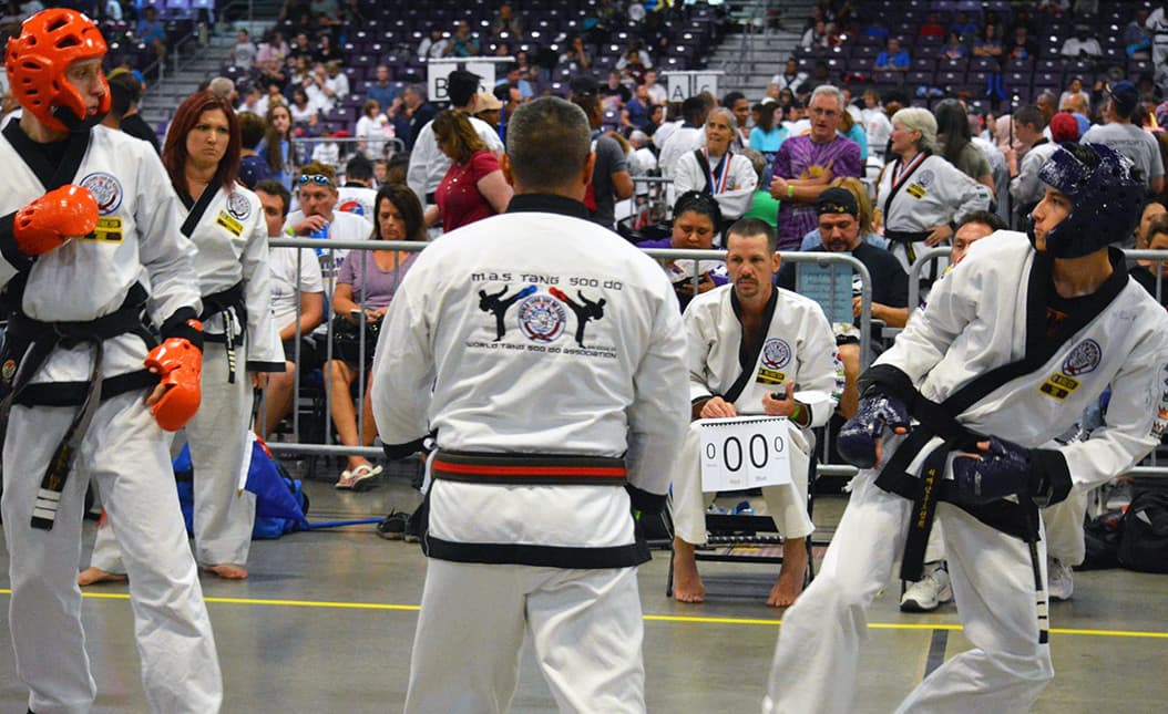 A center judge standing between two sparring competitors in headgear, facing toward the scorekeeper holding a 0-0 scoreboard.