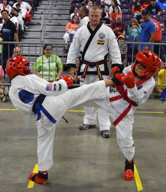 A center judge watching closely as two competitors execute a high kick exchange near head height.