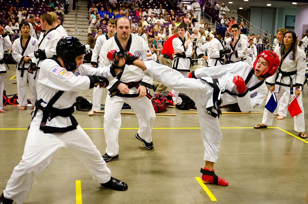Two competitors sparring mid-kick while corner judges holding red and blue flags watch closely from ringside.