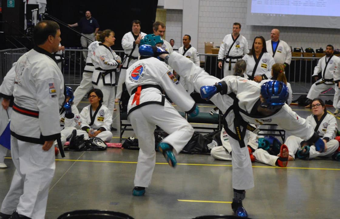 Two team members in protective sparring gear seated together at ringside, waiting for their match.