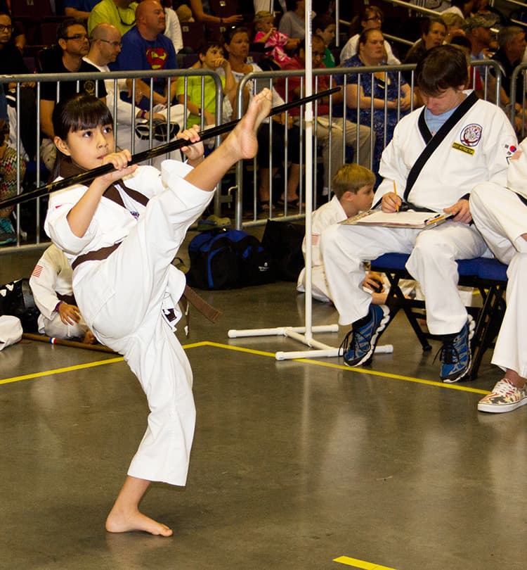 A young competitor performing a bong (staff) hyung while a ring official records notes beside the ring.